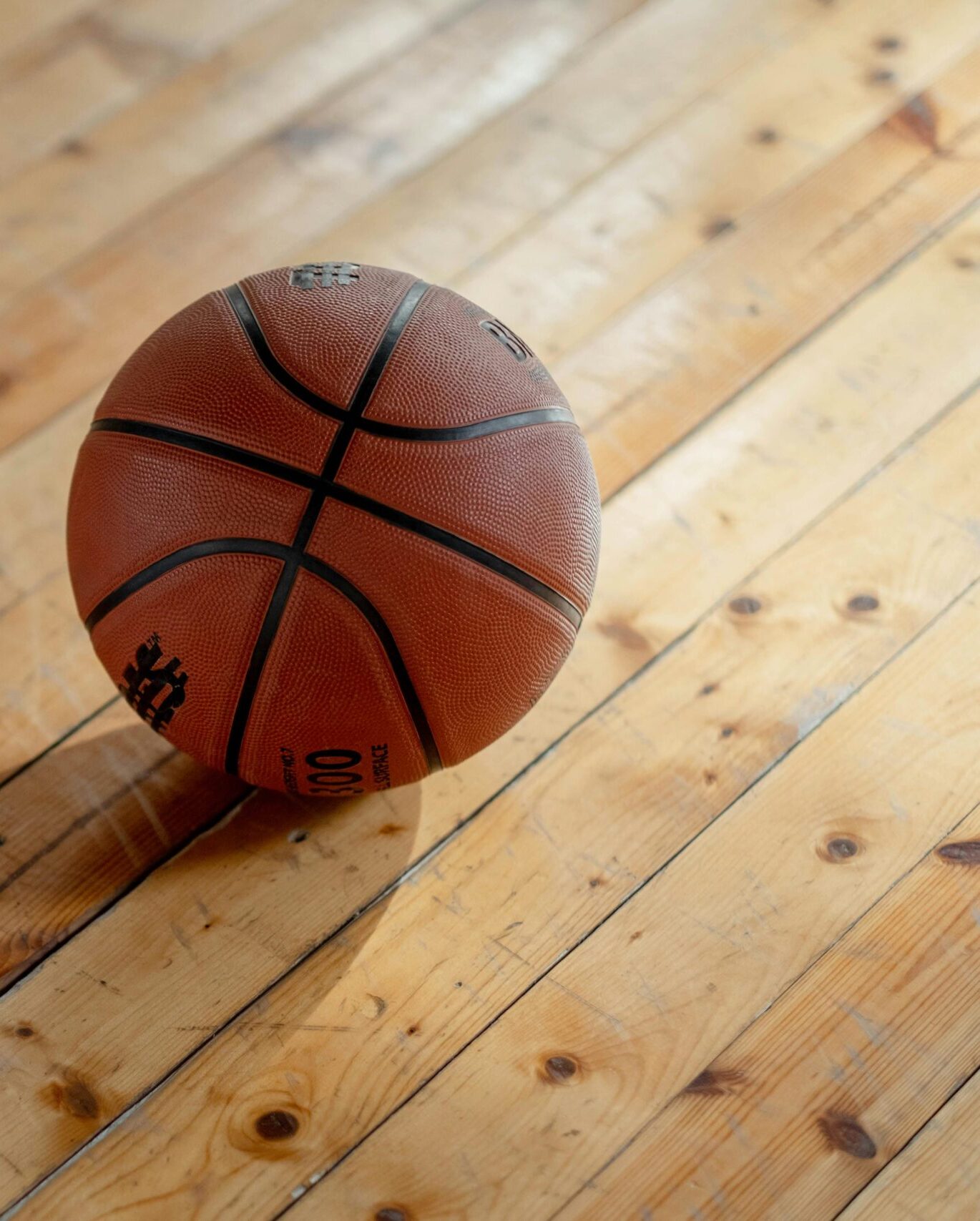 A single basketball resting on a polished wooden gym floor, casting a shadow.